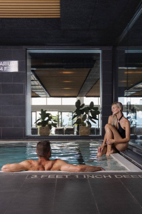Indoor pool setting with a man swimming and a woman sitting on the edge. Large windows in the background, plants visible. "3 Feet 6 Inch Deep" marked on the pool edge.