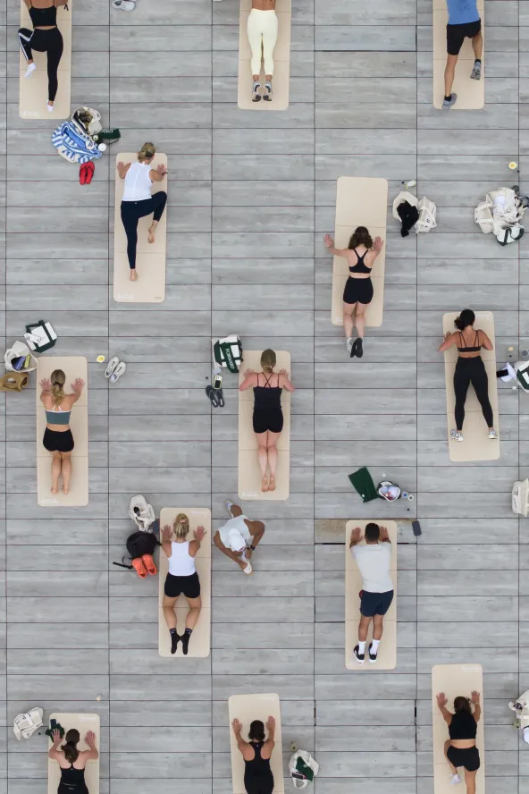 People lying on yoga mats in neat rows on a gray deck, viewed from above, some with bags beside them.