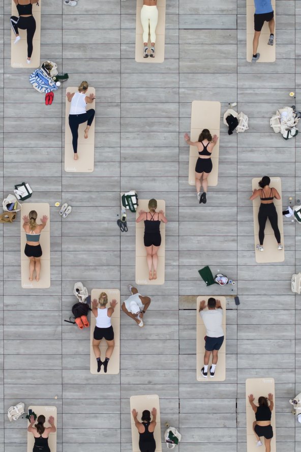 People lying on yoga mats in neat rows on a gray deck, viewed from above, some with bags beside them.