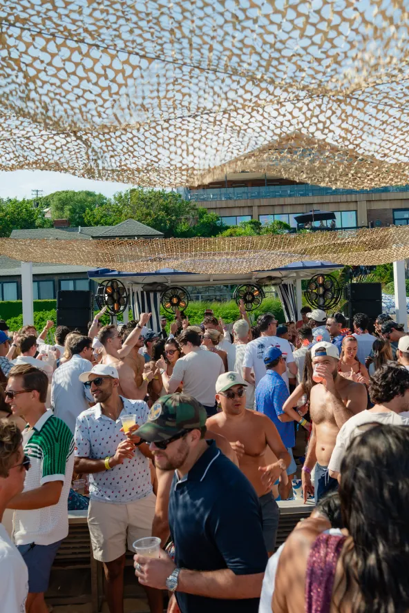 Crowded outdoor party with people mingling under woven canopies. Sunny day with greenery and buildings in the background.