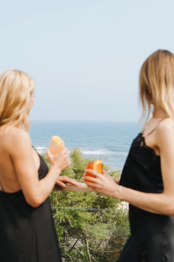 Two women in black dresses enjoy orange drinks on a balcony overlooking the ocean.