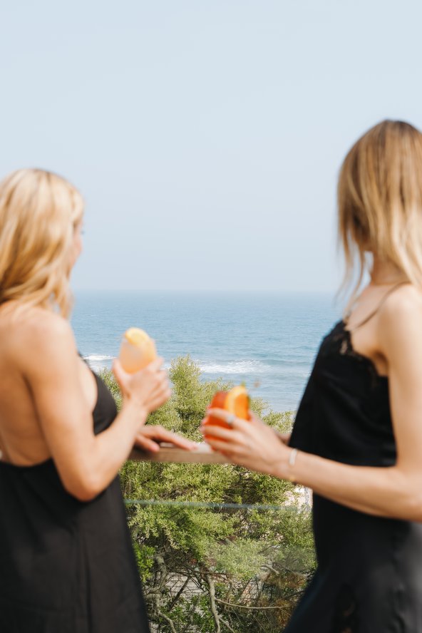 Two women in black dresses enjoy orange drinks on a balcony overlooking the ocean.