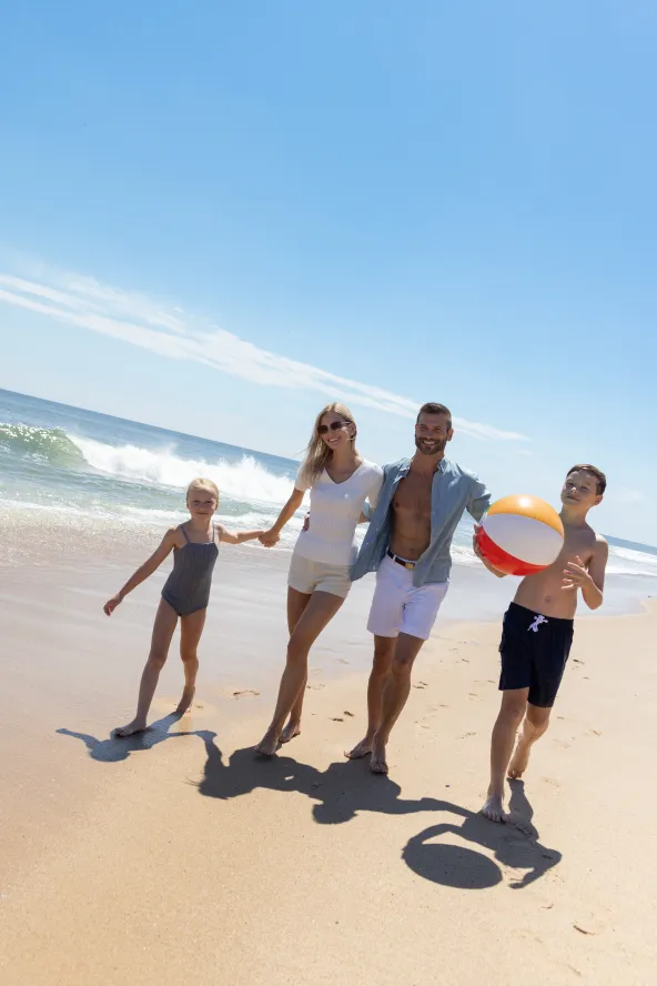 Family of four walks on sunny beach, child holding beach ball, ocean waves in background.