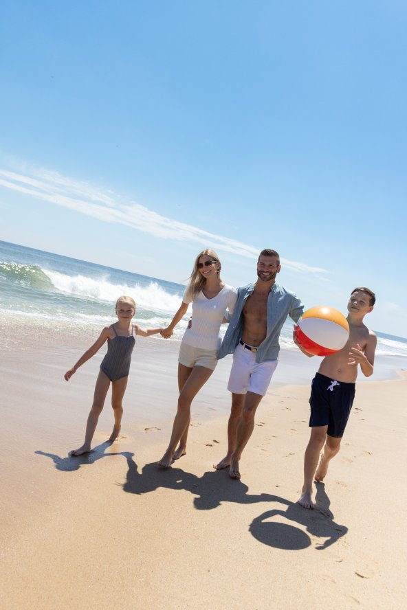 Family of four walks on sunny beach, child holding beach ball, ocean waves in background.