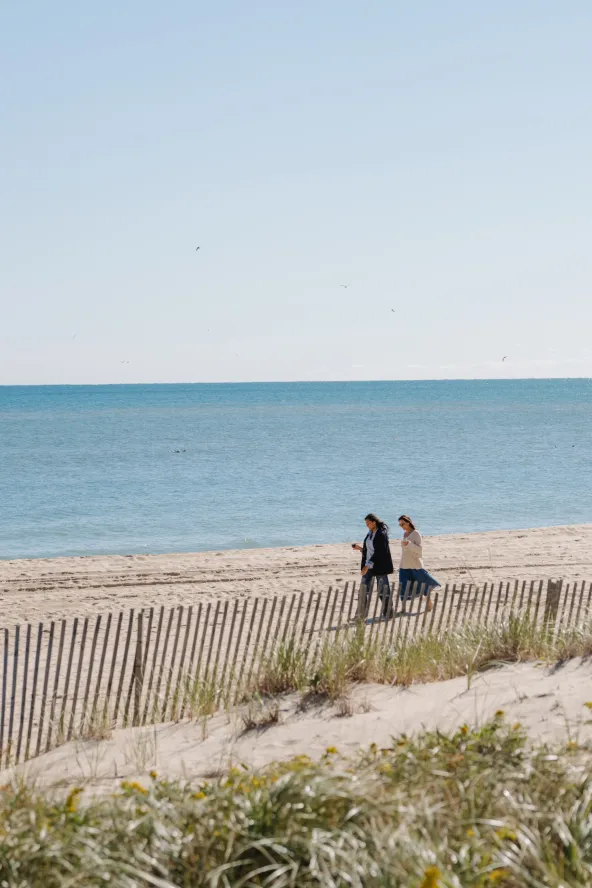 Two people walk along a sandy beach with a wooden fence, while the ocean stretches out under a clear blue sky.