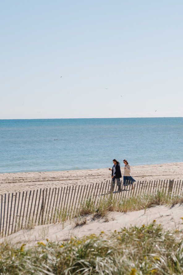 Two people walk along a sandy beach with a wooden fence, while the ocean stretches out under a clear blue sky.