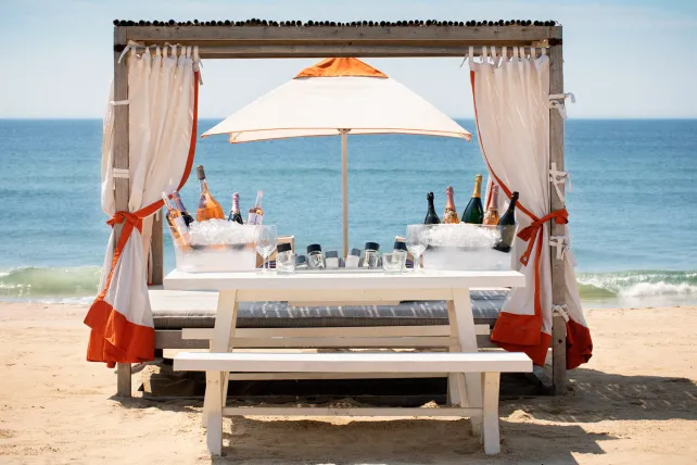 Beach cabana with a white table, drinks on ice, under a parasol, facing the ocean. Curtains tied with orange accents.