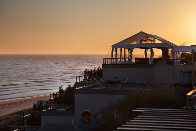 Sunset over a calm beach, with modern beach houses on the right. A bird flies in the sky, and the ocean reflects the warm orange light.