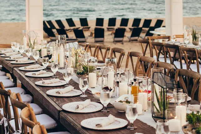 Elegant beachside dining setup with long tables, neatly arranged plates, glasses, and candles, overlooking a row of lounge chairs by the sea.