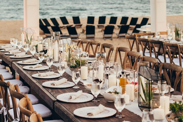 Elegant beachside dining setup with long tables, neatly arranged plates, glasses, and candles, overlooking a row of lounge chairs by the sea.