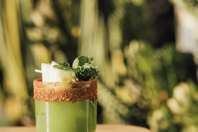 Green cocktail with a salted rim garnished with cucumber and herbs on a wooden table, blurred greenery in the background.