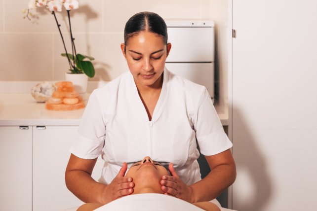 Female massage therapist giving facial to spa guest with flowers in background.