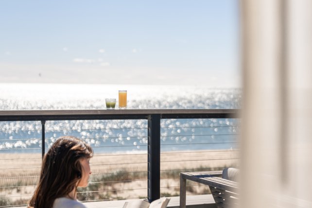 Woman reading on a balcony overlooking a sunny beach and ocean, with clear blue skies and sparkling water.