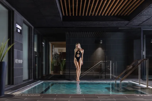 A woman in a black swimsuit stands by an indoor pool in a modern, dimly lit space with dark walls and wood ceiling accents.