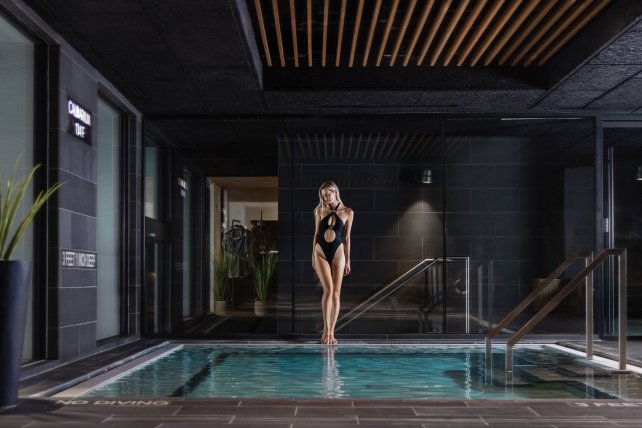 A woman in a black swimsuit stands by an indoor pool in a modern, dimly lit space with dark walls and wood ceiling accents.