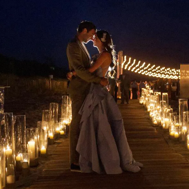 A couple embraces on a candlelit boardwalk at night, with string lights in the background.