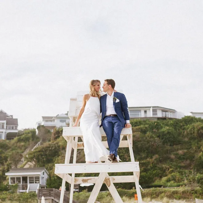 A couple in wedding attire sits on a lifeguard chair overlooking a beach, with houses and greenery in the background.
