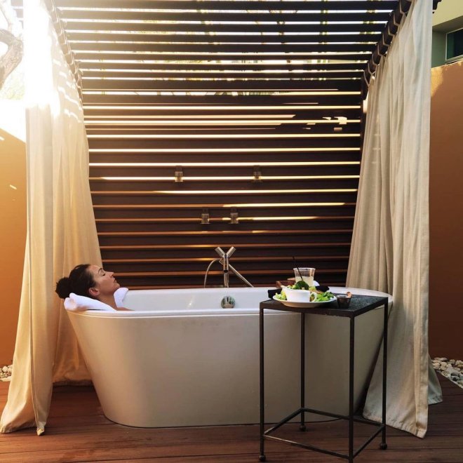 Woman relaxing in an outdoor bathtub under a wooden pergola, with a small side table holding a tea set.