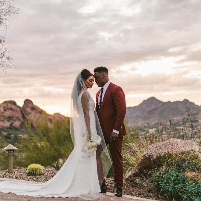 Bride in white gown and groom in burgundy suit stand close at sunset, with desert landscape and mountains in the background.