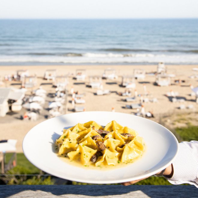 Pasta dish being presented by server with beach view in background.