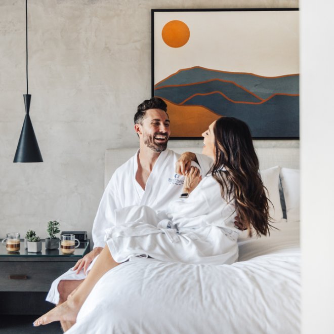 Couple lounging on the bed in robes sharing a moment with coffee on nightstand and desert artwork in background.