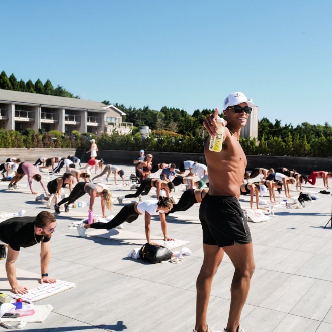 Outdoor fitness class on a sunny day, with a large group exercising and an instructor holding a beverage, wearing a hat.