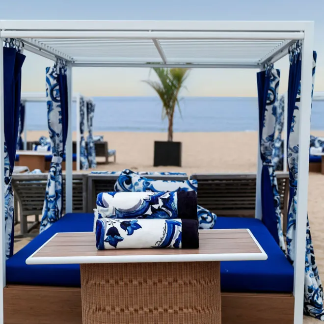 Beach cabana with blue and white decor on sandy shore, calm sea and sky in background, single palm tree visible in distance.