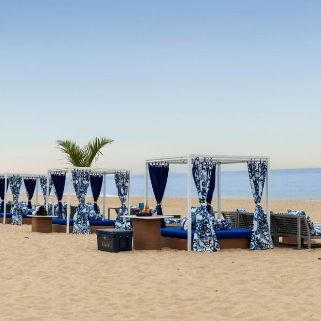 Cabanas with blue and white curtains on a sandy beach, palm trees nearby, calm sea under a clear sky.