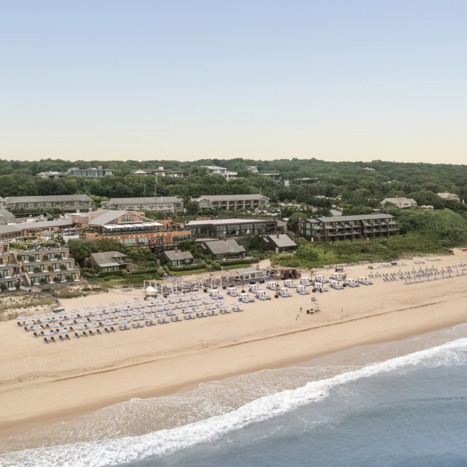 Aerial view of a beach with rows of umbrellas, buildings set back among green trees, and gentle waves on the sandy shore under a clear sky.