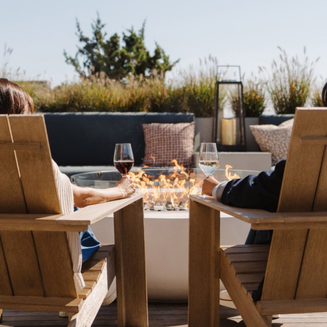 Two people relax in wooden chairs by a fire pit, holding glasses of wine on a sunny patio.