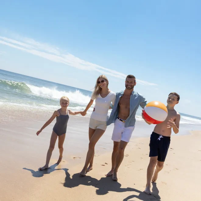 Family of four walks on sunny beach, child holding beach ball, ocean waves in background.
