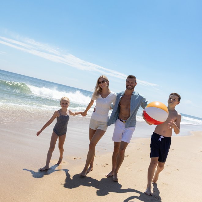 Family of four walks on sunny beach, child holding beach ball, ocean waves in background.