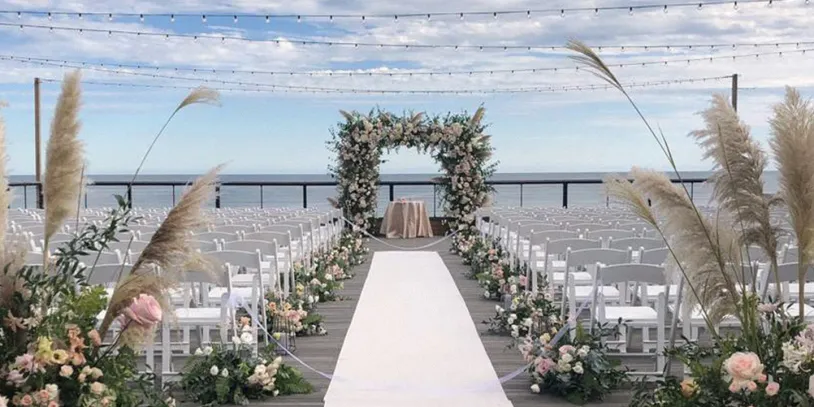 Outdoor wedding venue by the ocean, with white chairs, floral arch, and pampas grass lining the aisle under a blue sky.