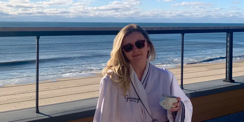 Woman in sunglasses and robe holding a cup, standing on a deck overlooking a calm ocean under a cloudy sky.
