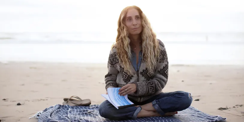 Person with long hair sitting cross-legged on a beach, holding a book, wearing a patterned sweater. Overcast sky and sea in the background.