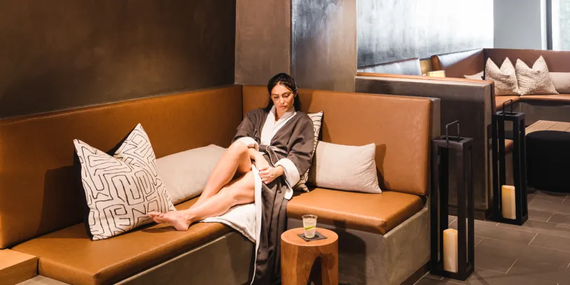 A woman in a spa robe sits on a leather bench with pillows, a wooden side table, and candle lanterns nearby.