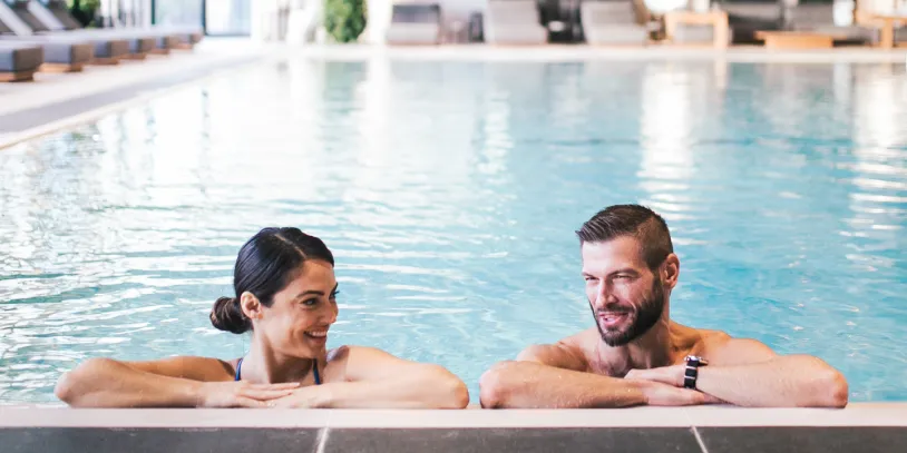 A man and woman relax in an indoor pool with lounge chairs in the background.