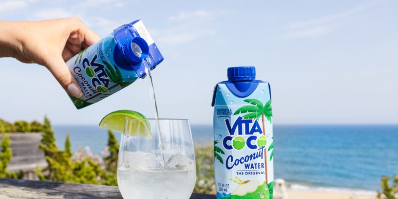 Coco Vita being poured into cocktail glass with ice and lime wedge with beach in backdrop.