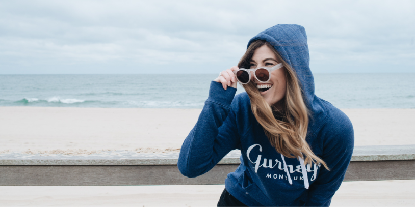 A woman in a blue Gurney's Resorts hoodie and sunglasses with the beach in the background