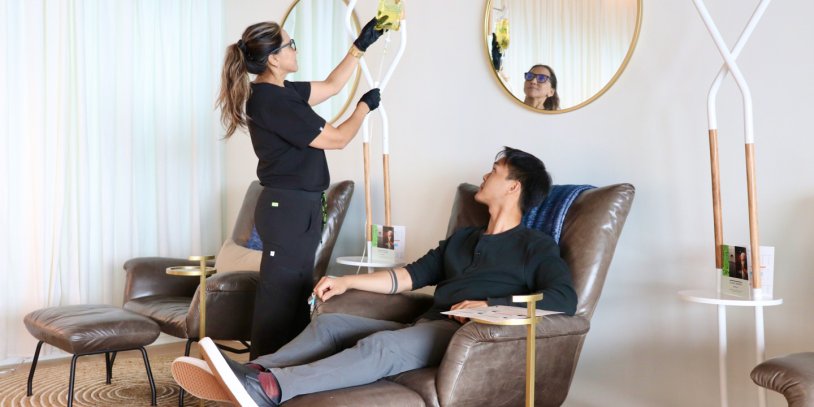 Woman administering IV Drip to guest in comfortable lounge chair in his hotel room.
