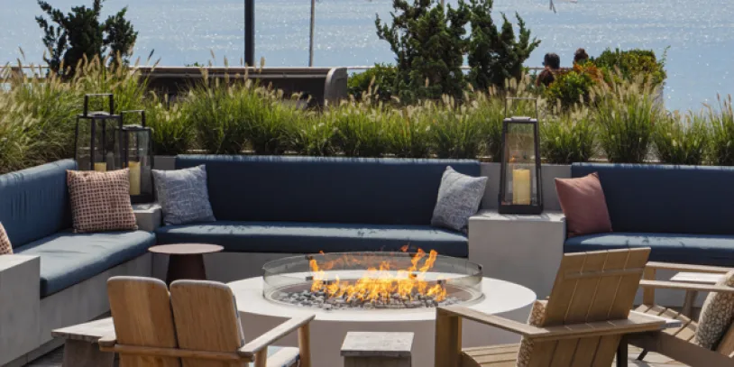 Cozy outdoor seating around a fire pit with ocean view, wooden chairs, and string lights overhead. Umbrellas and greenery enhance the relaxing atmosphere.
