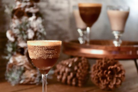 Golden brown holiday cocktail in sugar lined glass with Christmas tree and pine cones in background.