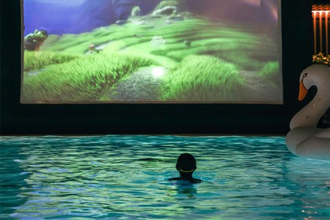 A child floats in an indoor pool in the dark facing a movie projected on a screen