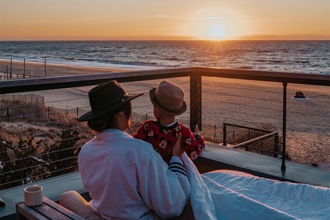 A woman and a small child sit under a blanket watching the sunrise on a deck overlooking the ocean.