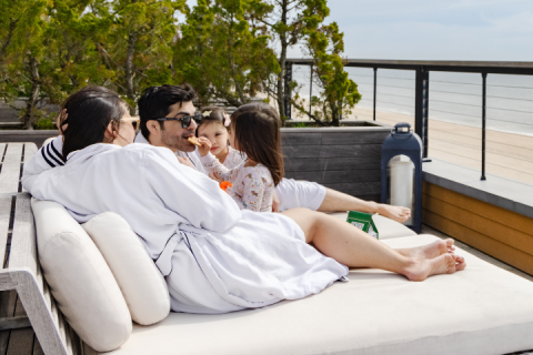 A family of four in robes looks out onto the ocean while the daughter feeds her father a cookie