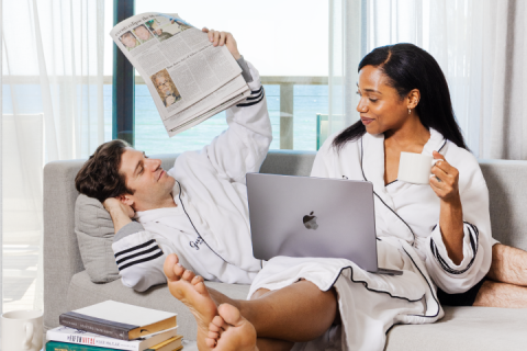 A couple in robes read the newspaper and browse on a laptop with the ocean in the background