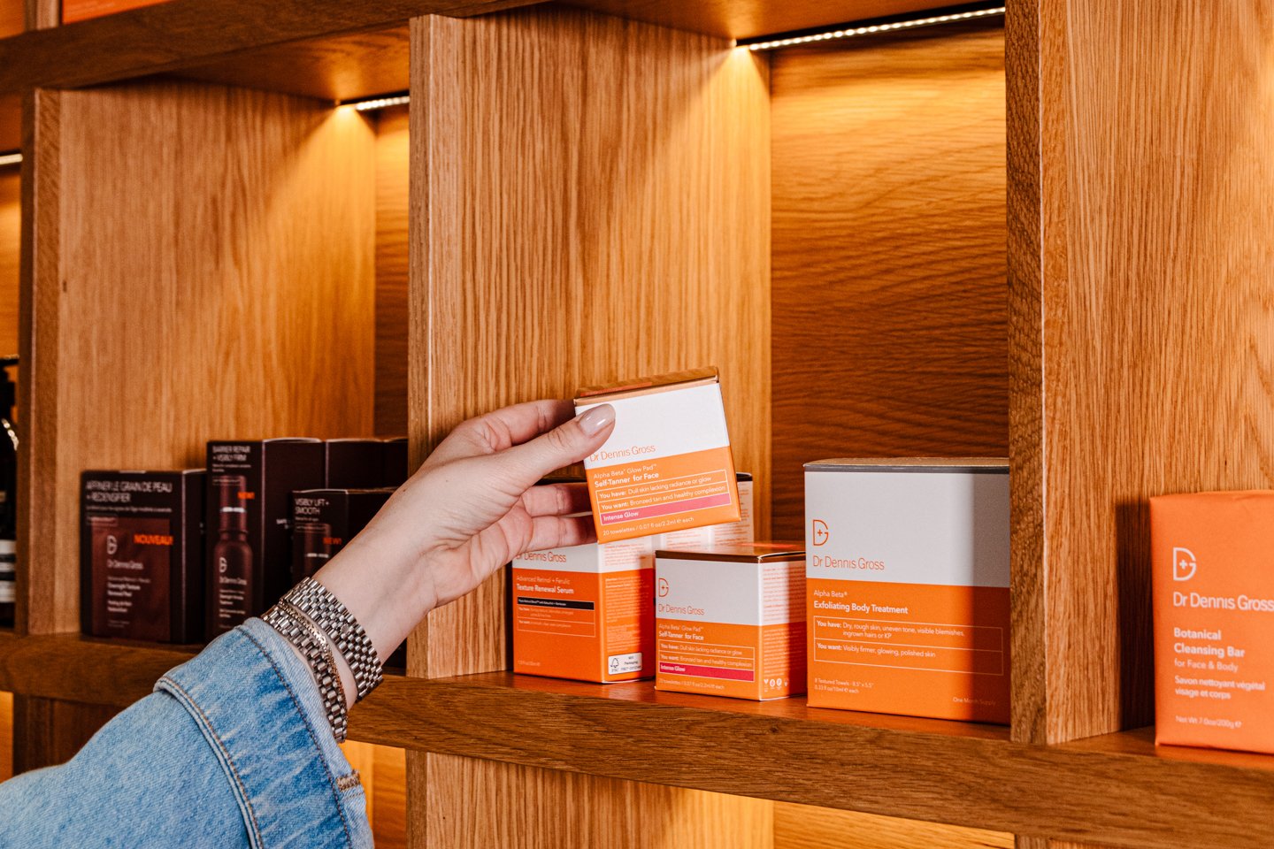 Woman in denim long sleeve picking up Dr. Dennis Gross product from wood shelf in orange and white packaging.