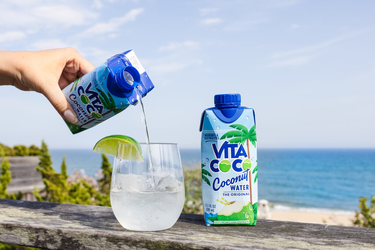 Coco Vita being poured into cocktail glass with ice and lime wedge with beach in backdrop.