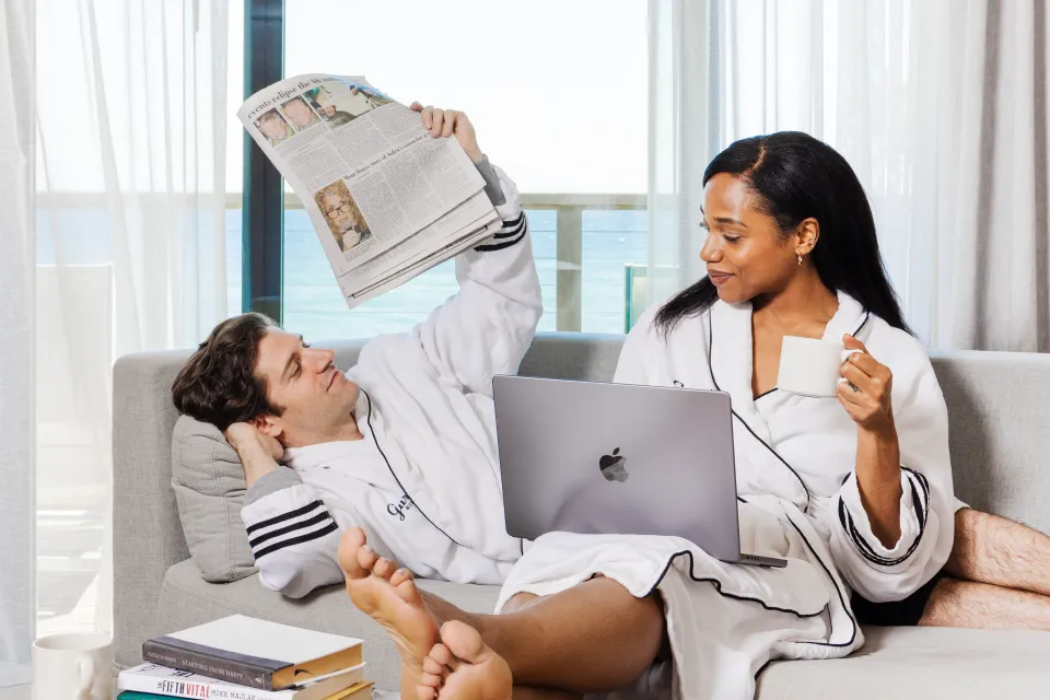 A man and woman in robes sitting on a sofa in a lobby lounge area with a laptop, newspaper, and coffee.