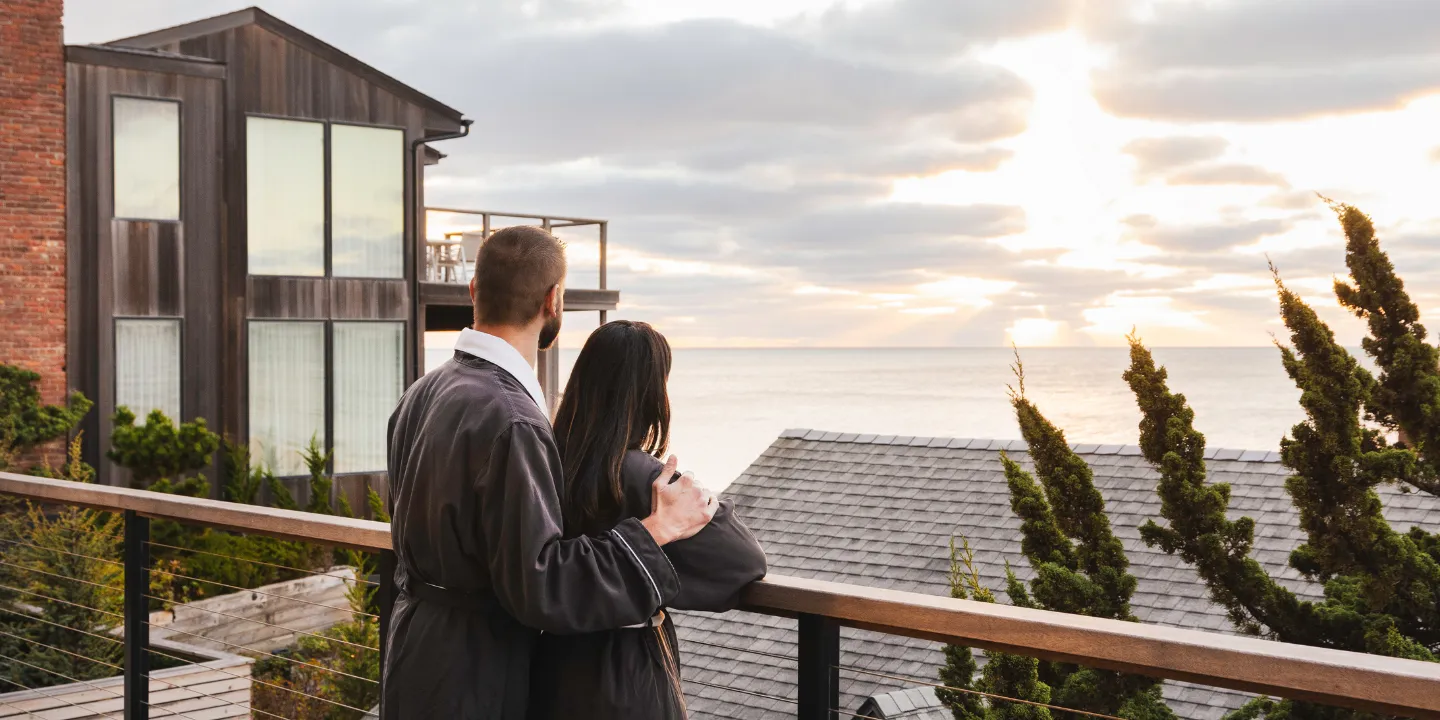 A couple in robes stands at a railing overlooking the ocean at sunset.